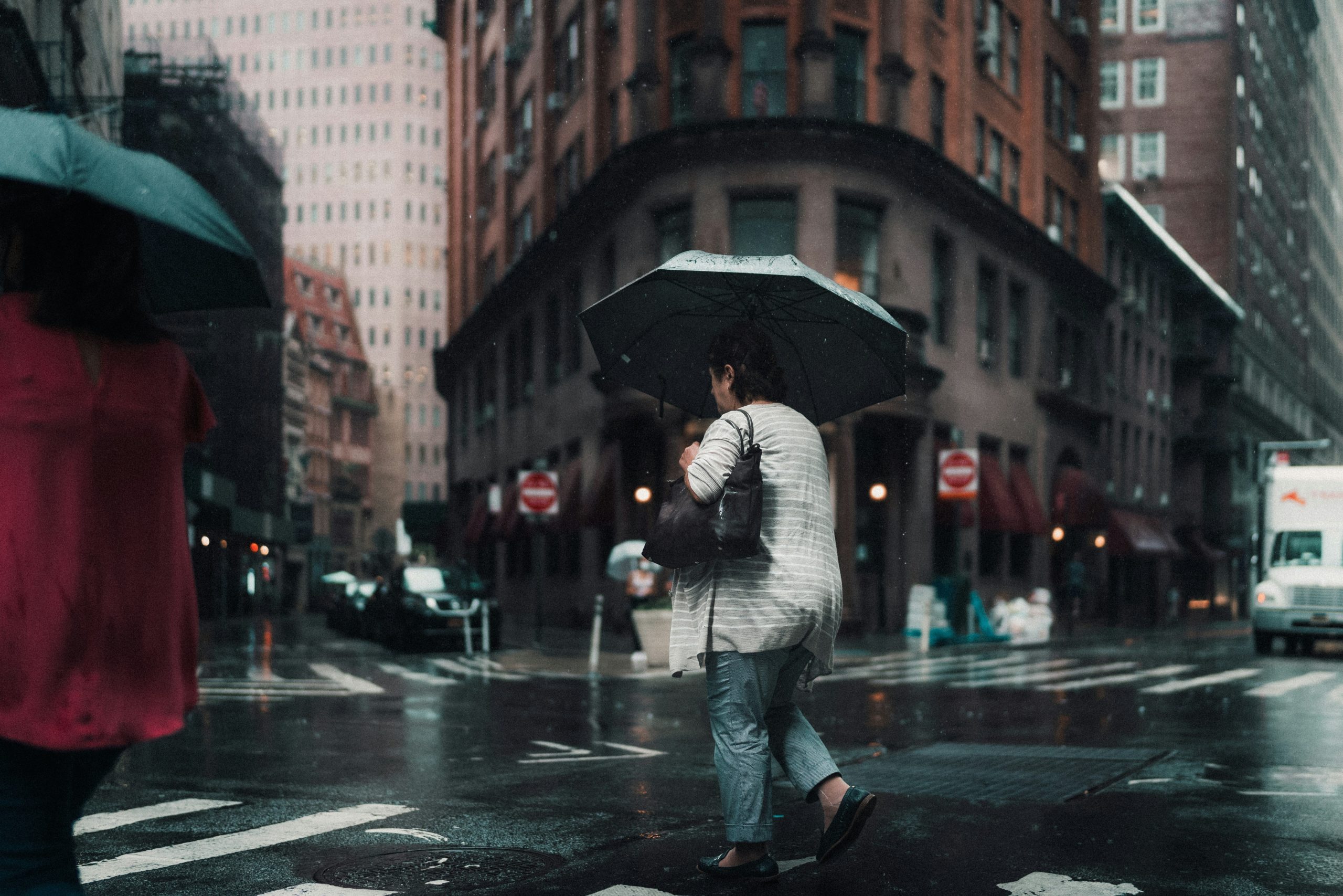 Urban travelers walking through heavy rain while one wears a reflective blue rain cover on their backpack.